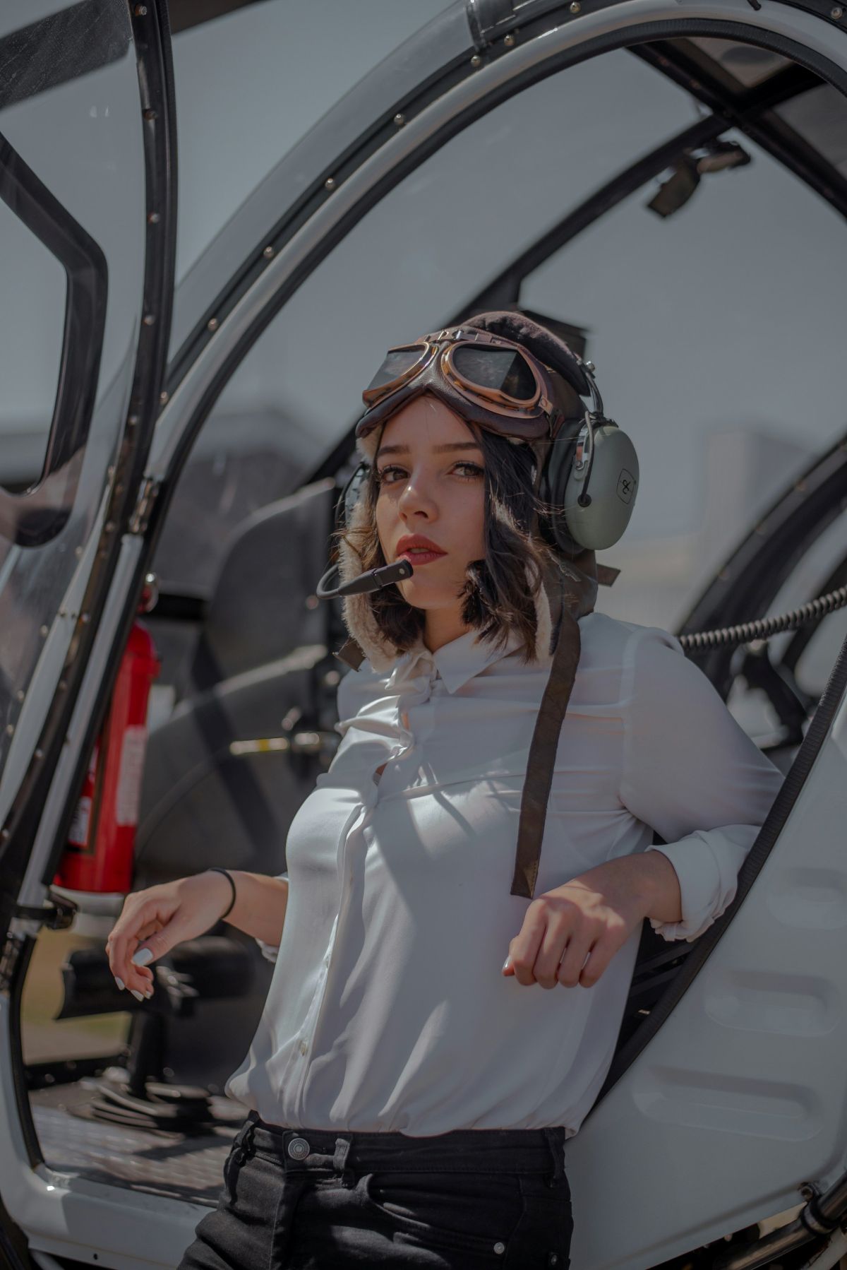  Female pilot in aviation headset and vintage goggles standing beside aircraft cockpit.